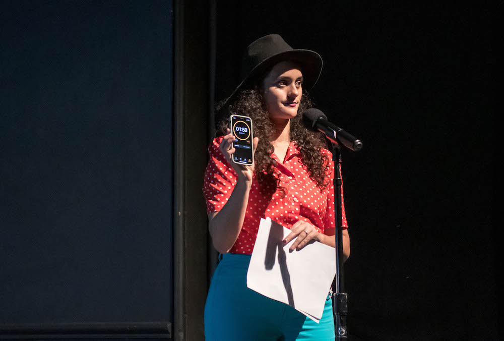 a brown skinned woman with curly long dark hair and a black cap. stands at  mic . in one hand she holds an iphonewith a timer that reads 1:58 in the other hand she holds white papers... she is wearing a red blouse with white polka dots and light blue jeans 